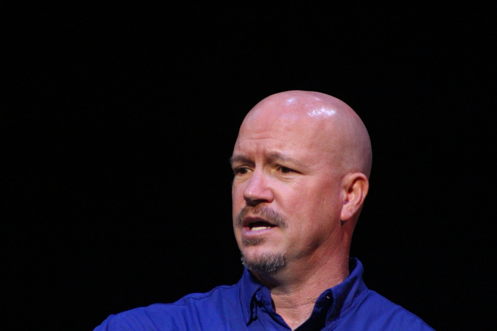 A bald man in a blue shirt speaking into a microphone.