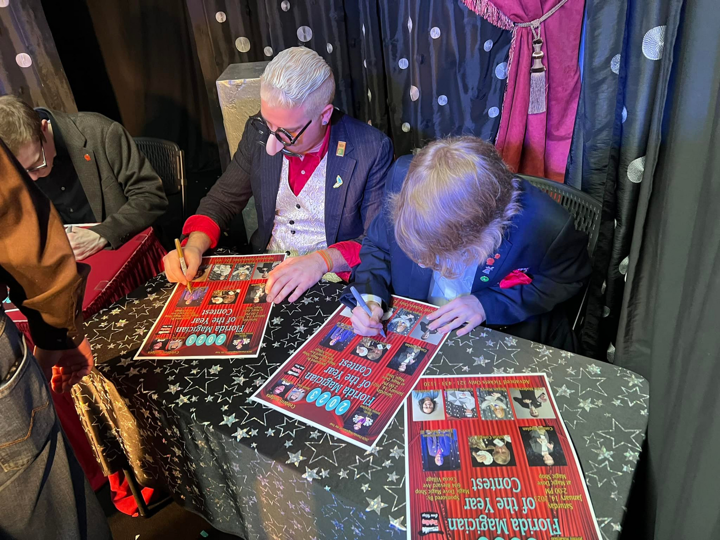 Two men signing posters at a table in front of a red curtain.